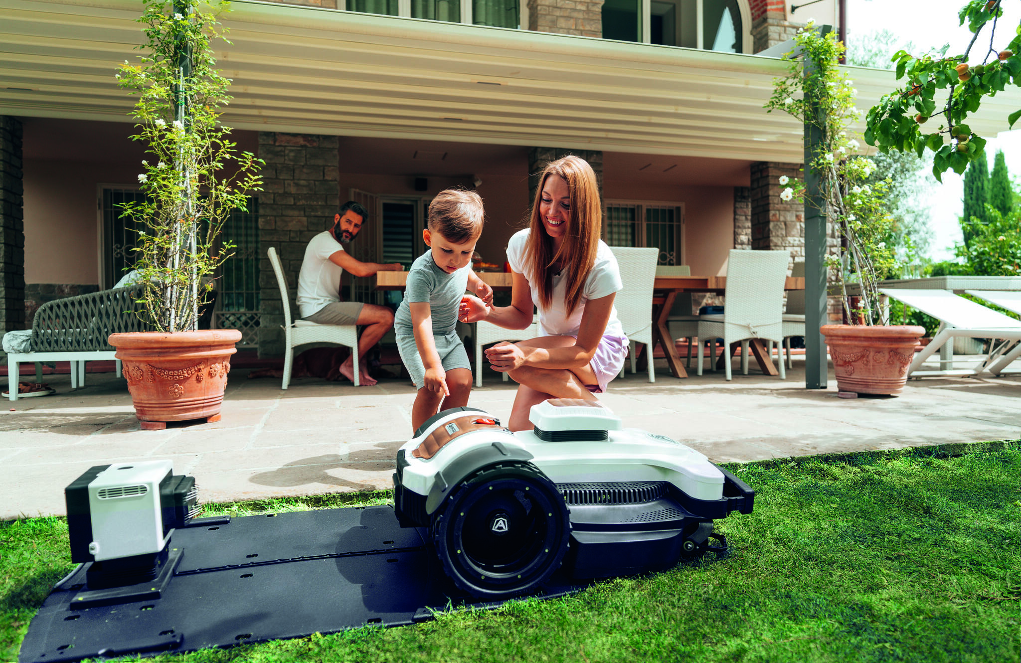 Robot koszący na trawie przed domem. Matka z małym dzieckiem i ojciec w tle